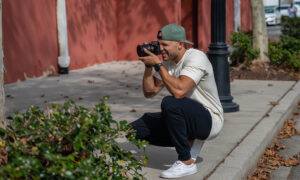 RBV Media A man crouches on a sidewalk, cap snug on his head, snapping photos with precision for RBV Media. In the background, a red building and lush plants add a splash of color to this Myrtle Beach scene. Myrtle Beach, Content, Ads, Web, Social, Email