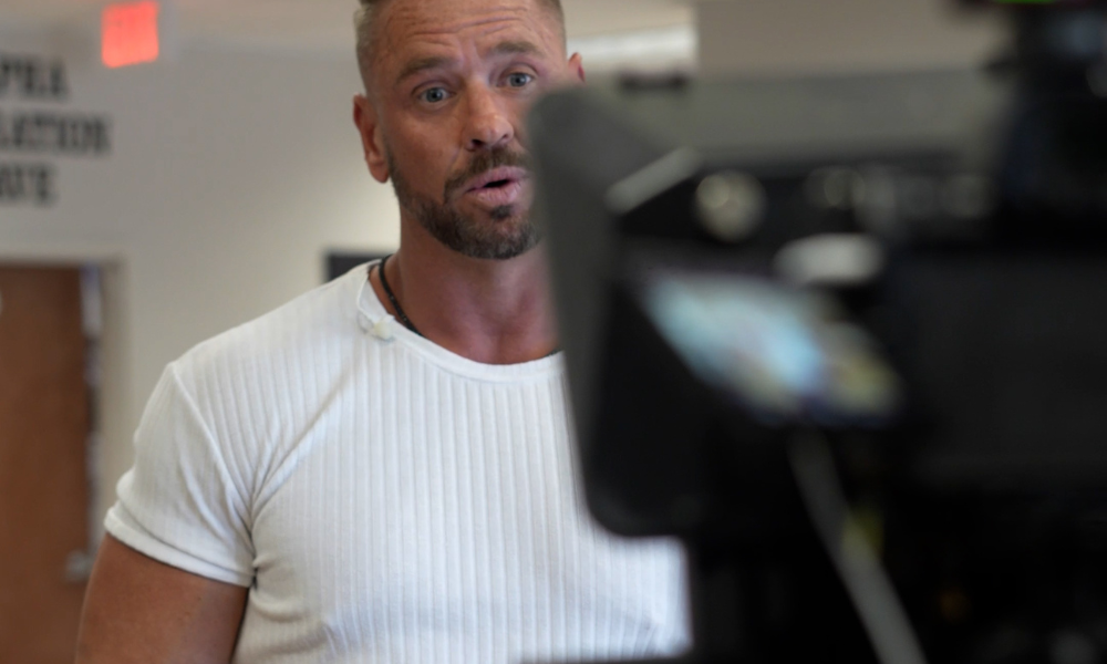 RBV Media A man in a white ribbed shirt stands indoors, looking toward a camera with a surprised expression. A blurred camera setup is visible in the foreground. Myrtle Beach, Content, Ads, Web, Social, Email