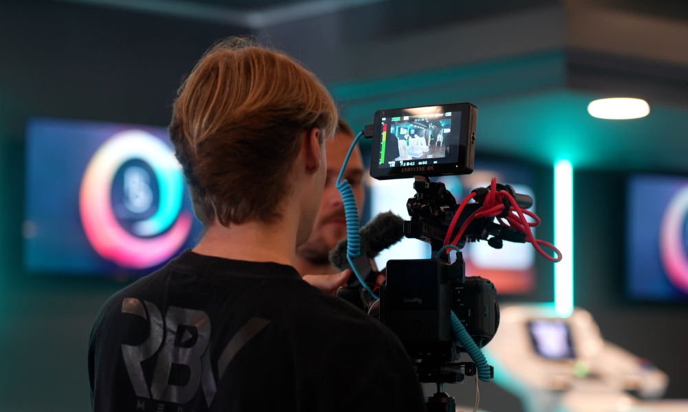 RBV Media Person with light brown hair operates a professional video camera, capturing footage in a modern studio with screens and neon lighting in the background. Myrtle Beach, Content, Ads, Web, Social, Email