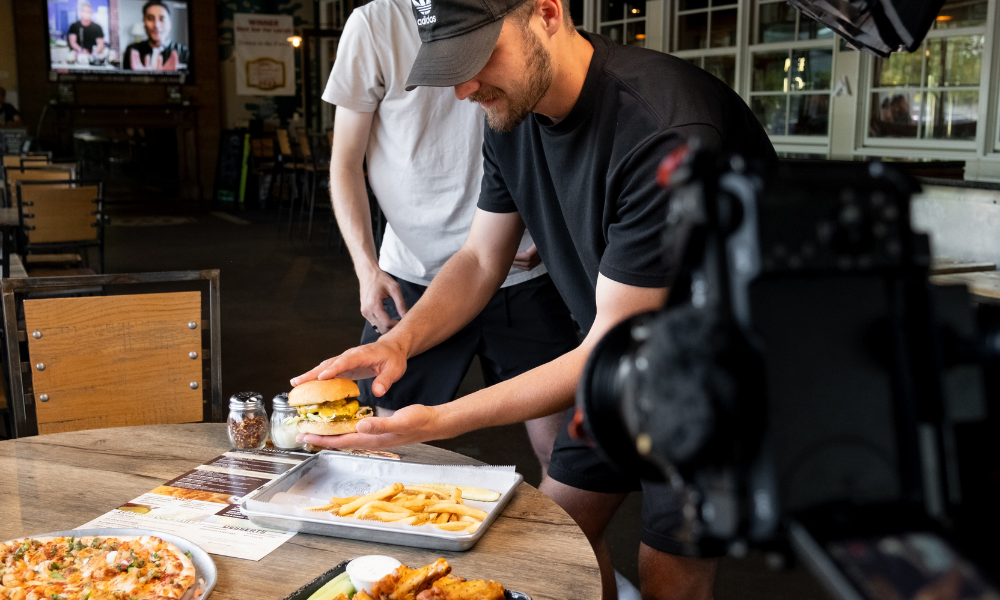 RBV Media Two people at a restaurant prepare to eat, with one holding a burger over a tray of fries. A camera is set up nearby to record the meal, and pizza is also on the table. Myrtle Beach, Content, Ads, Web, Social, Email