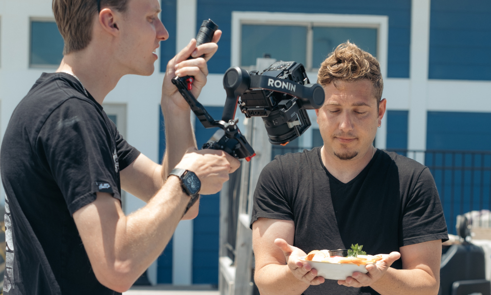 RBV Media Two men outdoors; one films with a stabilized camera while the other holds a plate of food, eyes closed, both wearing black shirts. Myrtle Beach, Content, Ads, Web, Social, Email